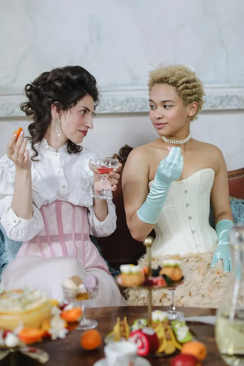 Two women in period costumes enjoy an elegant afternoon tea with assorted treats and drinks.